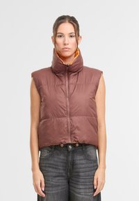 Vesta typu puffer, hnedý vonkajší materiál, oranžový vnútorný materiál, vysoký golier, bez rukávov, zapínanie na zips, nastaviteľný spodný lem s gombíkmi, hladká textúra.