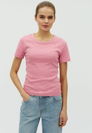 Nuori nainen, jolla on hiukset kiinni ja joka pukeutunut vaaleanpunaiseen lyhythihaiseen toppiin ja vaaleansinisiin farkkuihin, seisoo käsi taskussa valkoista taustaa vasten.