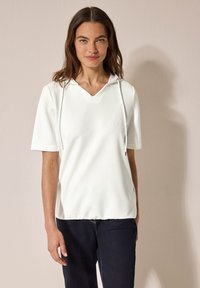 Haut blanc à manches courtes avec capuche et décollet�é en V, présentant un tissu texturé. Le design est décontracté, associé à un jean foncé.