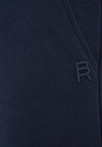 Temno modri bombažni joggerji z teksturirano tkanino, stranskimi žepi in gumiranem logotipu "R" na spodnjem delu noge.