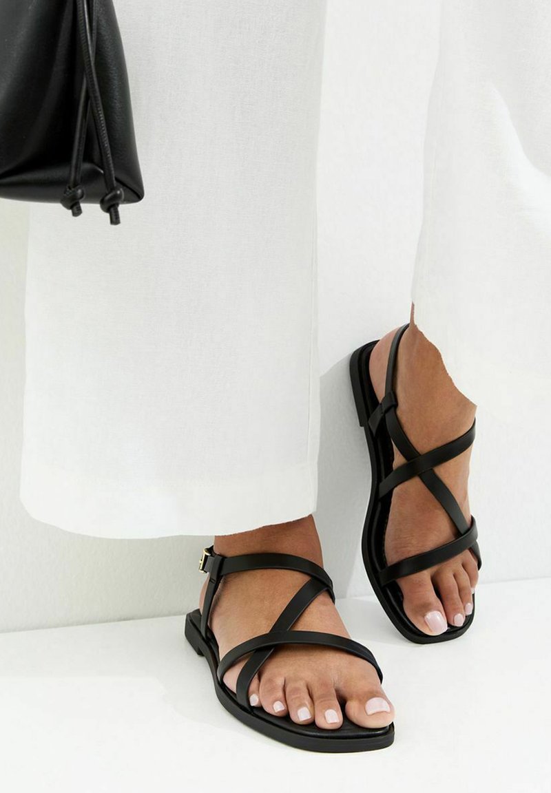Pies con sandalias planas negras de tiras combinadas con pantalones blancos de pierna ancha y sosteniendo un bolso de mano de cuero negro.