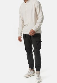 Mann in einem off-white Langarm-Henley-Shirt, schwarzen Cargo-Hosen mit mehreren Taschen und beigefarbenen weißen Sportschuhen, stehend.