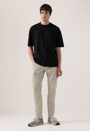 Mladý muž stojaci s rukami v vreckách, oblečený v čiernej košeli s krátkym rukávom, béžových cargo nohaviciach a sivých teniskách na svetlom pozadí.