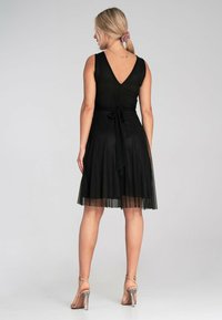 Schwarzes ärmelloses Kleid mit einem V-Ausschnitt hinten, Bindegürtel in der Taille, fließendem Rock und einem durchsichtigen Überzug. Kombiniert mit transparenten Riemchen-Highheels.