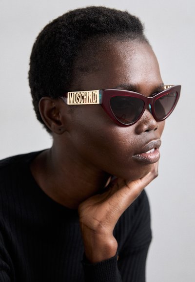 Kattöga-solglasögon med mörkröda bågar, guldakcenter och Moschino-logotypen på bågarna. Visas på en modell med kort hår.