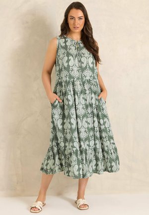 Kvinde står med hænderne i lommerne iført ærmeløs grøn midi-kjole med hvidt blomstermønster og hvide slip-on sandaler.
