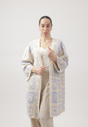 Kvinde iført lang beige og lyseblå hæklet cardigan over hvid top og beige bukser, stående mod en ensfarvet lys baggrund.