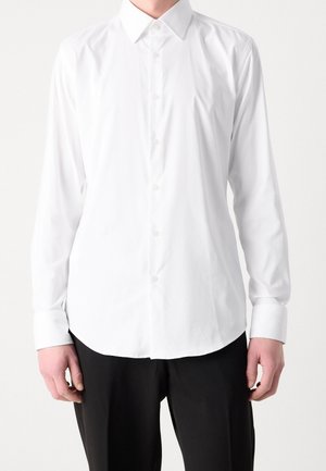 Person trägt ein langärmeliges weißes Hemd mit Knöpfen und schwarze Hose, steht vor einem schlichten, hellen Hintergrund.