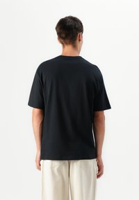 T-shirt de algodão preto com decote redondo e mangas curtas, apresentado pela parte de trás, combinado com calças de tom claro. Textura suave.
