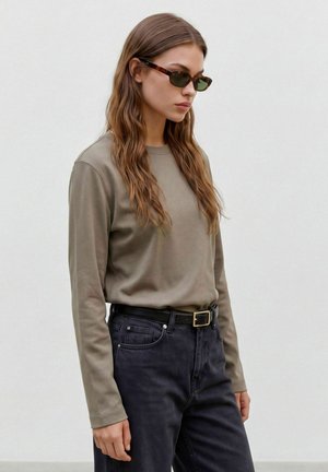 Femme aux longs cheveux ondulés portant des lunettes de soleil foncées, une chemise à manches longues vert olive, un jean noir et une ceinture noire, debout de côté contre un mur blanc.