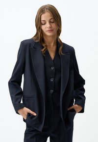 Blazer su misura blu navy con rever a punta, tasche laterali e vestibilità rilassata, abbinato a un gilet coordinato con bottoni rotondi.