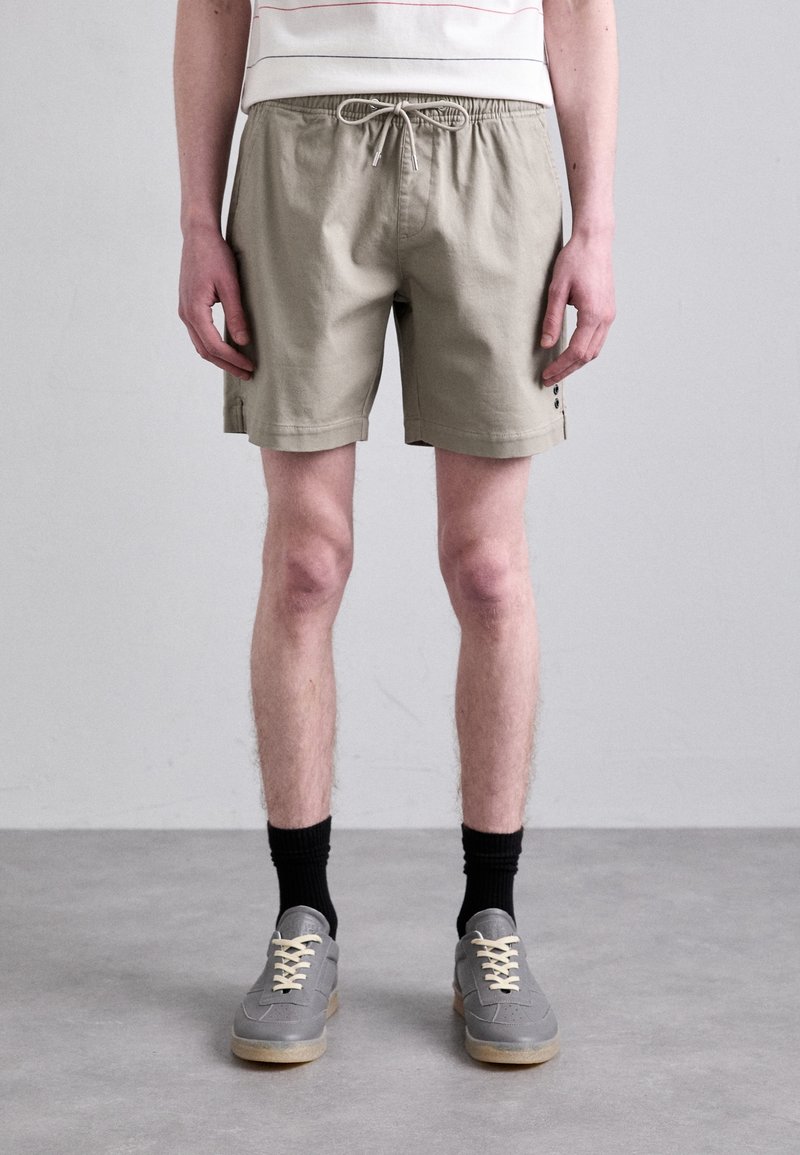 Personne portant un short beige avec cordon, des chaussettes noires et des chaussures gris à lacets, debout sur un sol gris devant un mur gris uni.