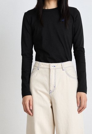Personne portant une chemise noire à manches longues rentrée dans un pantalon large taille haute couleur crème avec des coutures visibles.