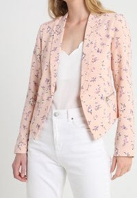 Femme portant un blazer floral rose pâle sur un haut blanc à bord festonné et un pantalon blanc taille haute, debout les mains détendues.