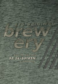 Zelená texturovaná tkanina s reliéfním textem: "220 ORIGINAL pivovar" a geometrickými liniemi a souřadnicemi níže.