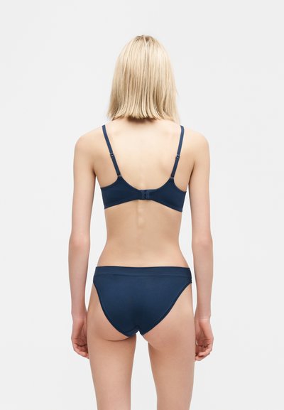 Γυναίκα με ξανθά μαλλιά, φοράει μπλε σουτιέν και εσώρουχο που ταιριάζει, στεκόμενη πλάτη προς το φακό σε απλό λευκό φόντο.