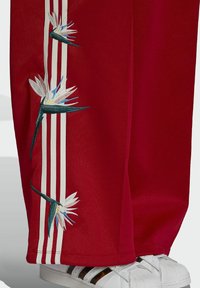 Red sweatpants with white and black stripes, featuring embroidered white and blue flowers along the side, paired with white sneakers.