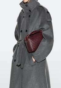 Person trägt einen grauen, doppelreihigen Mantel mit Gürtel und hält eine weinrote, gewebte Ledertasche unter dem Arm.