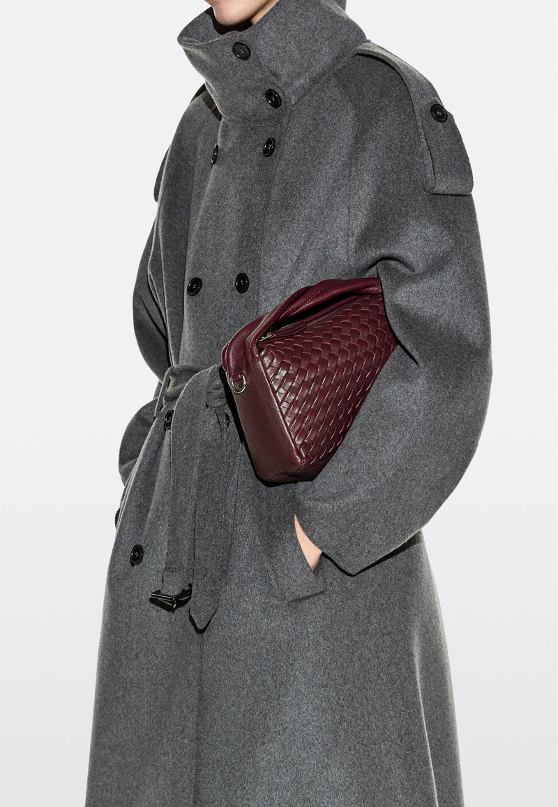 Person trägt einen grauen, doppelreihigen Mantel mit Gürtel und hält eine weinrote, gewebte Ledertasche unter dem Arm.