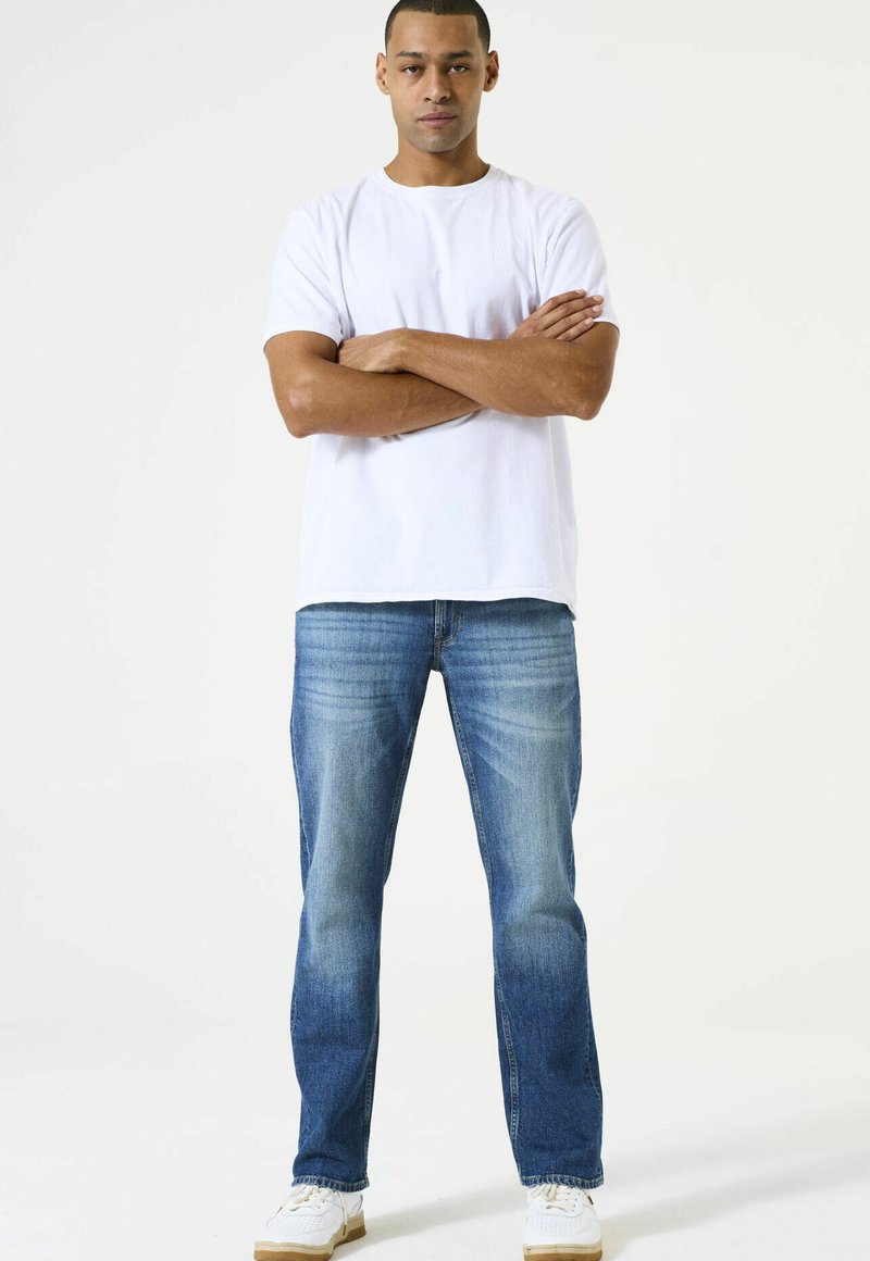 Witte t-shirt met korte mouwen, denim jeans in lichtblauw met vervaagde details, en witte sneakers met een bruine zool. Model staat met gekruiste armen.