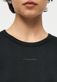 Κοντινό πλάνο του κάτω μέρους του προσώπου και του λαιμού ενός ατόμου που φοράει ένα μαύρο ριμπ μπλουζάκι Calvin Klein με στρογγυλή λαιμόκοψη.