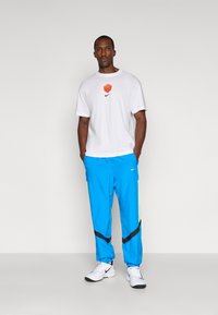 White short-sleeve t-shirt with an orange logo, paired with bright blue pants featuring black and lime green accents, and white sneakers.