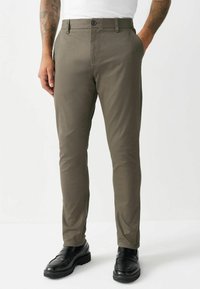 Olive green slim-fit chinos made from smooth fabric, featuring a button closure and side pockets; paired with black loafers.
