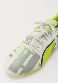 Zapato de fútbol con un estampado geométrico en gris y blanco con acentos en amarillo brillante. Hecho de material sintético, cuenta con malla texturizada y cordones.