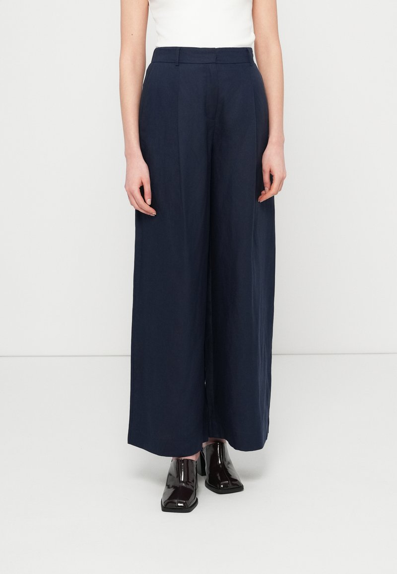 Μπλε σκούρο παντελόνι φαρδιάς γραμμής από ελαφρύ ύφασμα, με ψηλή μέση, πιέτες και λεία υφή, συνδυασμένο με γυαλιστερά μαύρα παπούτσια.