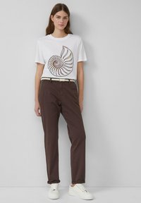 T-shirt blanc avec un graphique de coquille de nautile colorée, associé à un pantalon marron et des baskets blanches. Le pantalon est doté d'une ceinture tissée.
