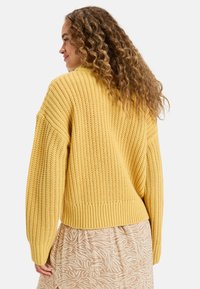 Pull en laine jaune avec une coupe décontractée, des poignets et un ourlet côtelés. Présente un tissage texturé et un dos court. Associé à une jupe à motifs.