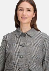 Młoda kobieta z prostymi brązowymi włosami, ubrana w szary w kratkę płaszcz zapinany na guziki z kołnierzem i przednimi kieszeniami, na białym tle.