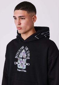Felpa nera con tasca frontale, presenta una grafica floreale e un logo, cappuccio con cordini e dettagli bianchi sulle maniche. Texture del tessuto morbida.