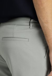 Pantaloni grigio chiaro con texture, dotati di una tasca posteriore e passanti per la cintura, parzialmente coperti da una maglia scura indossata da una persona.