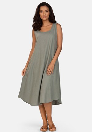 Kobieta o ciemnych włosach, ubrana w luźną, oliwkowozieloną sukienkę midi bez rękawów i brązowe klapki wsuwane, stojąca na jednolitym tle.