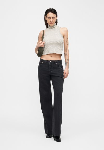 Femme tatouée portant un col roulé sans manches à rayures, un jean noir à jambes larges, des bottes noires, tenant un sac à bandoulière vert sur un fond blanc.