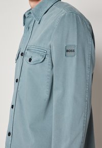 Homme portant une chemise bleu clair à boutons avec deux poches poitrine et un écusson logo "BOSS" sur la partie supérieure de la manche.