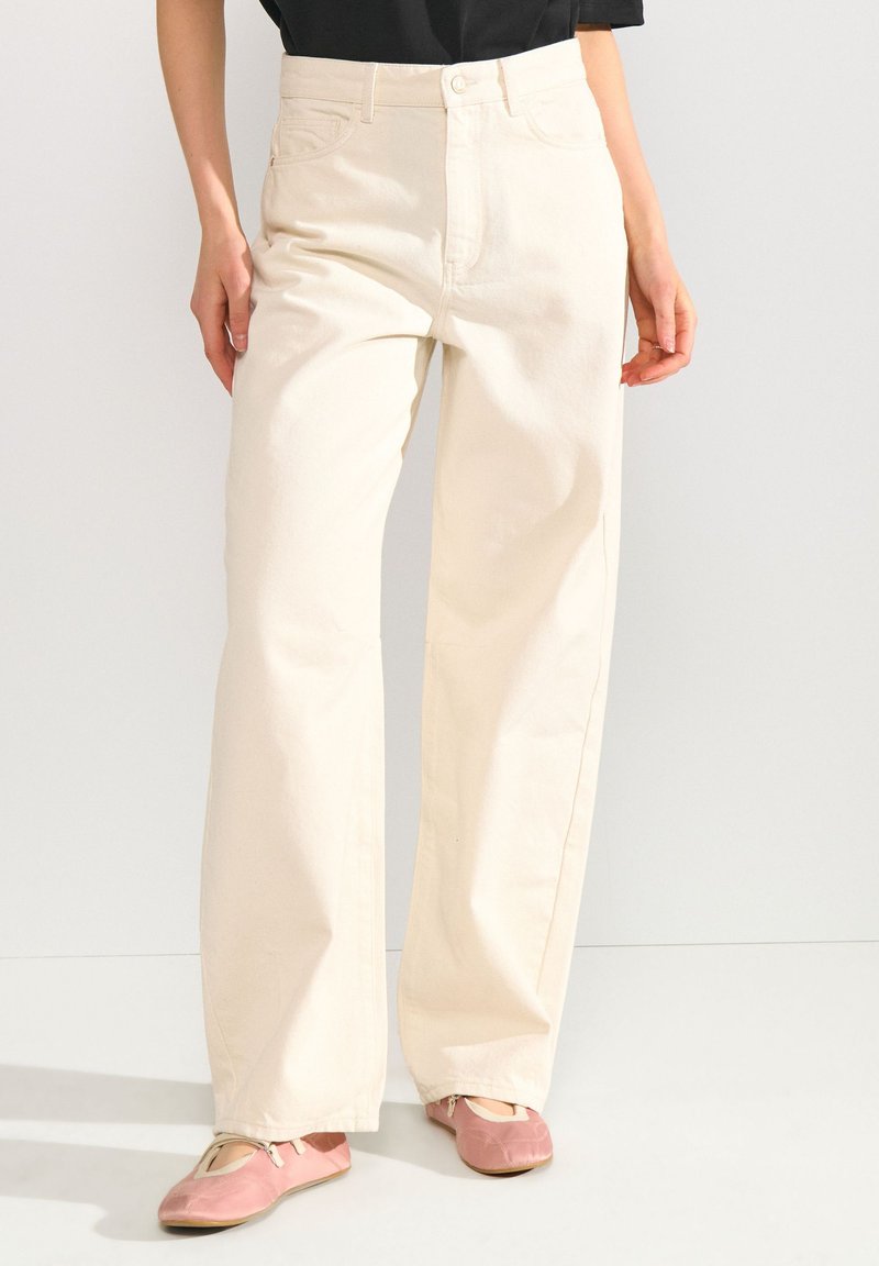 Femme portant un pantalon large écru, un haut noir et des ballerines roses, debout devant un fond clair uni.