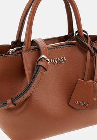 Brown textured leather handbag with dual handles, adjustable shoulder strap, zipper closure, and metal Guess logo on front and tag.