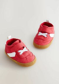 Zapatillas de tela rojas con correas de Velcro y cordones blancos, que cuentan con una suela de goma marrón texturizada y un detalle blanco en cada lado.