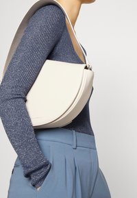 Sac à main en cuir couleur crème avec une forme incurvée, une sangle réglable et une texture lisse. Porté avec un haut bleu texturé et un pantalon tailleur.