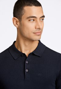 Tummansininen polo-paita, joka on valmistettu teksturoidusta kankaasta, sisältää klassisen kauluksen, kaksinappisen etukappaleen ja huomaamattoman brodeeratun logon rinnassa.