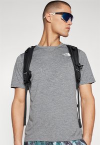 Camiseta gris de manga corta con un logotipo, tirantes de mochila negros y gafas de sol reflectantes azules, complementadas con pantalones cortos estampados.
