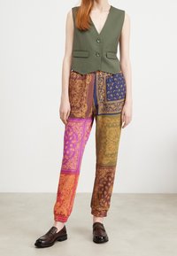 Chaleco sin mangas de color verde oliva con dos botones, combinado con pantalones de colores variados con patrones de paisley en púrpura, naranja y marrón.