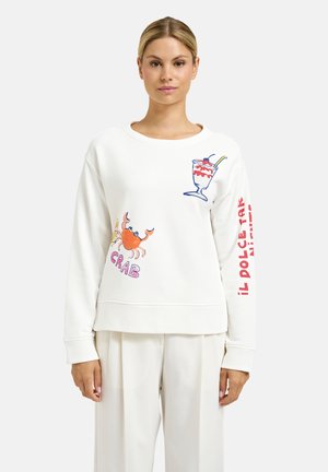 Frau trägt ein weißes Sweatshirt mit Illustrationen einer Krabbe und eines Dessertglases sowie roten Text auf dem rechten Ärmel, kombiniert mit weißen Hosen.