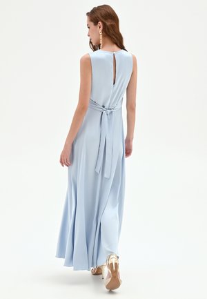 Femme portant une robe longue sans manches bleu clair avec une ceinture nouée et un dos en forme de trou de serrure, marchant de dos sur fond blanc.