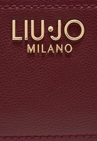 Burgunderrødt lædermateriale med en tekstureret overflade, der har et præget guld "LIU·JO MILANO" logo og kontrasterende lyserød søm.