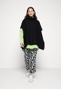 Svart strikket poncho med høy krage over en lys grønn skjorte, sammen med svarte bukser med blomstermønster og hvite joggesko.