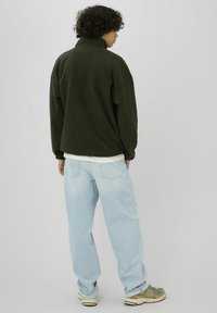 Pull-over en polaire vert foncé avec col montant et poignets côtelés, associé à un jean large bleu clair et des baskets grises, vu de dos.