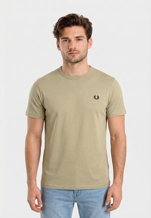 Jeune homme aux cheveux châtain clair portant un t-shirt beige uni avec un petit logo noir sur le côté gauche de la poitrine et un jean bleu clair.